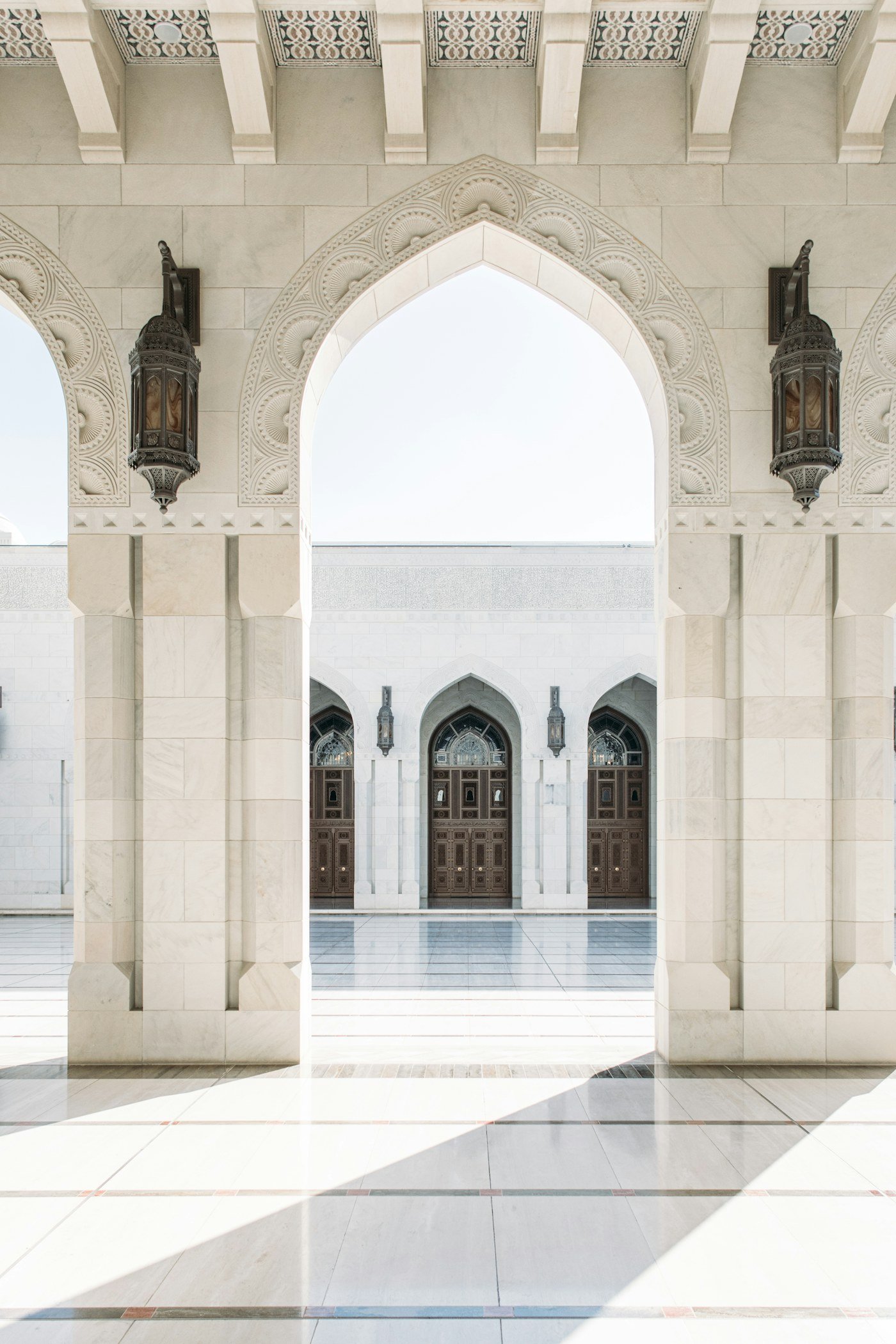 White marble arch with two lanterns, opening onto a sunlit mosque courtyard