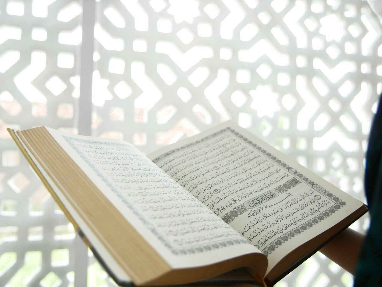 An open Quran held before a white geometric lattice window